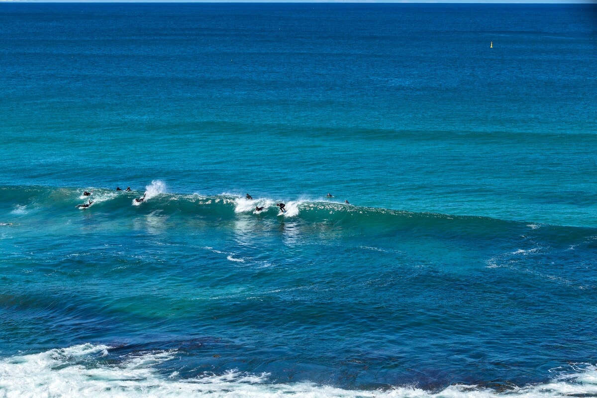 The image captures a vibrant seascape, featuring surfers riding the waves of Bondi Beach. The clear turquoise water contrasts with the white foam created by the surf. The horizon is open and unobstructed, illustrating the vastness of the ocean under a bright sky.
