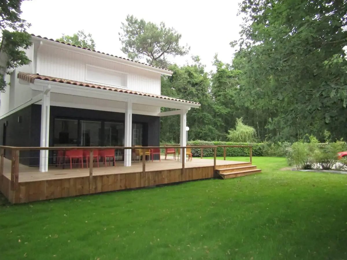 A spacious house with a wooden deck features multiple steps leading to the entrance. Large glass doors provide a view of the open interior. The surrounding area is landscaped with green grass and trees, contributing to a serene environment.
