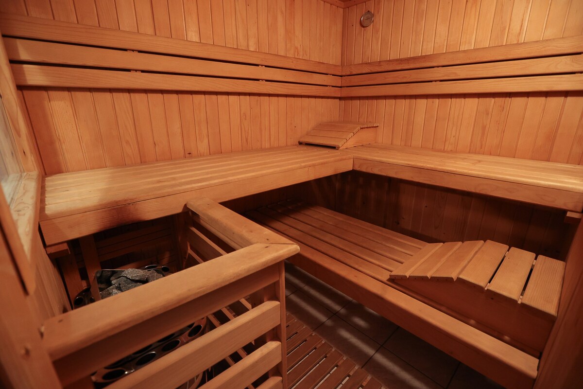 A wooden sauna interior is shown, featuring a spacious seating area with benches arranged along the walls. The wooden structure includes a traditional sauna heater, enhancing the warm, natural aesthetic of the space. Soft lighting creates a relaxed atmosphere.