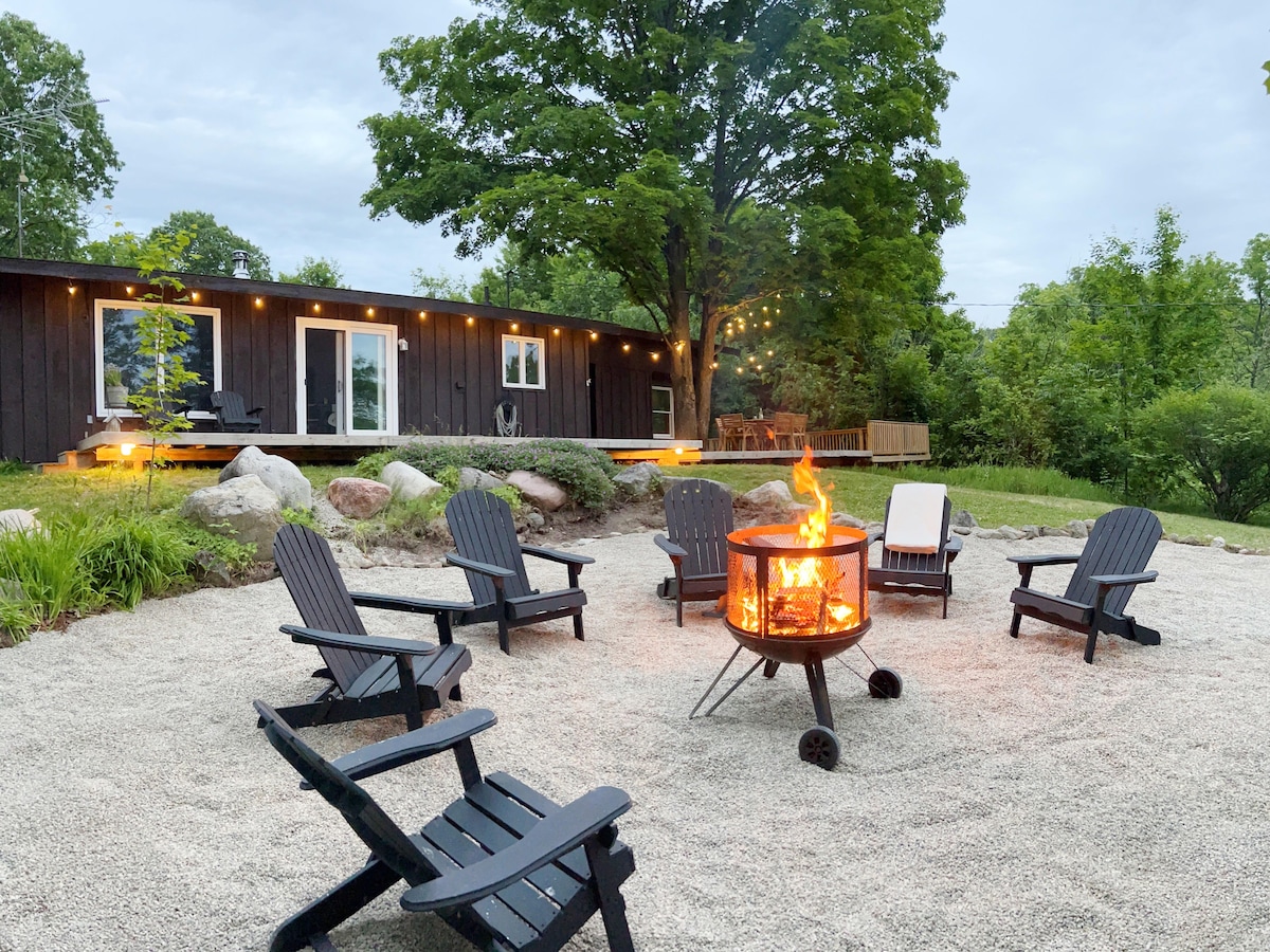 An outdoor fire pit surrounded by multiple black Adirondack chairs is situated on a sandy area. Soft evening light from string lights enhances the ambiance, while a rustic cottage is visible in the background, framed by greenery and tall trees.