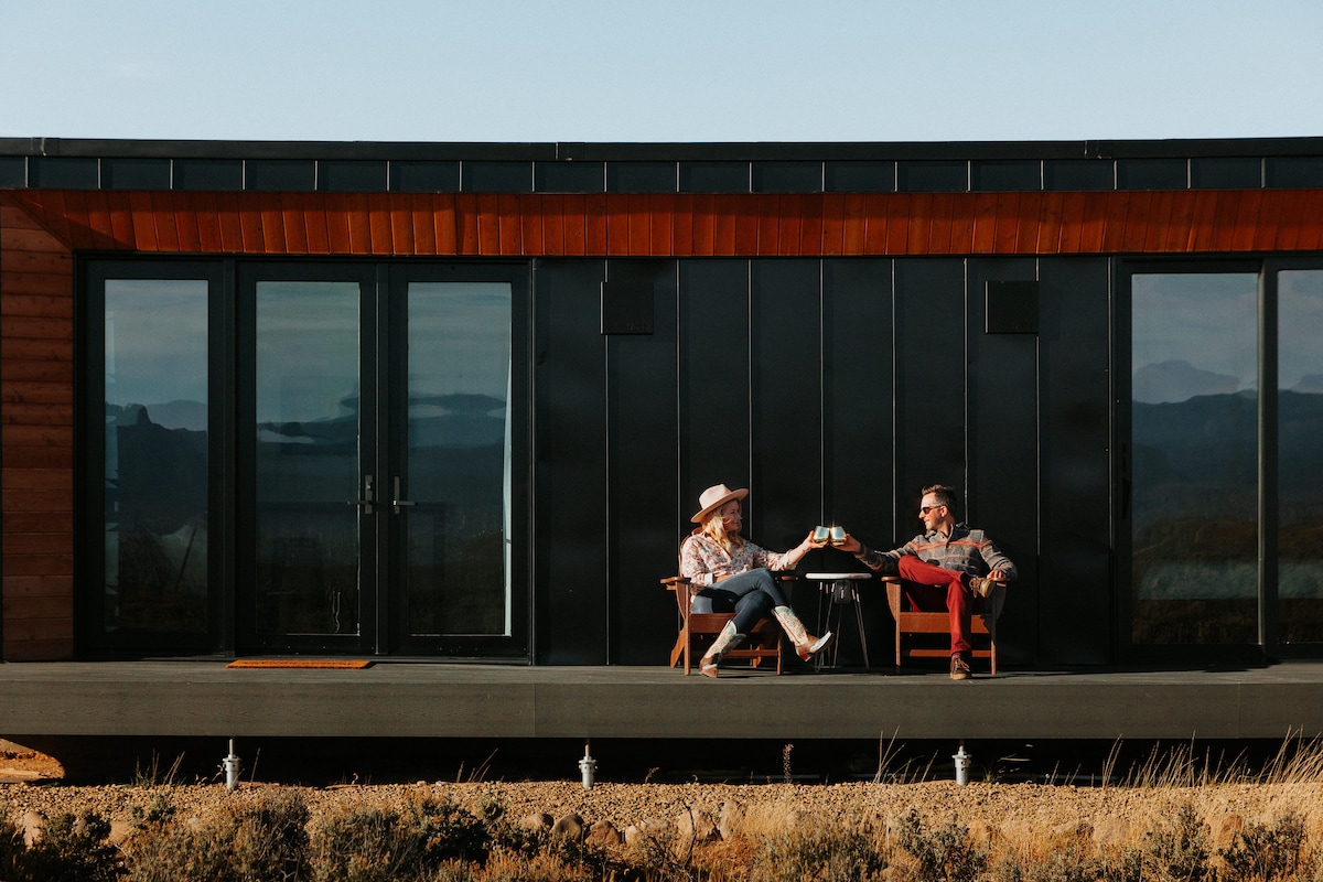A modern retreat features two individuals seated at a small table on a spacious deck. Large glass doors create a connection to the indoor space, while the high desert landscape stretches out in the background, highlighting the serene atmosphere of the setting.