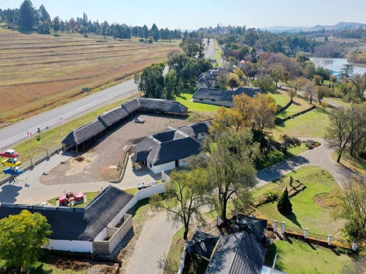 An aerial view of the Loch Athlone Estate reveals well-maintained grounds and several structures with thatched roofs. A road borders the estate, while fields and trees extend into the distance. The nearby lake is visible, enhancing the natural setting.