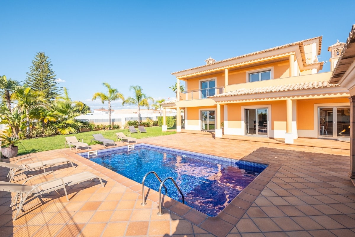 A spacious villa features a private pool surrounded by lounge chairs, set against a backdrop of palm trees and a clear blue sky. Large glass doors connect the indoor and outdoor spaces, allowing for effortless access to the sunlit terrace.