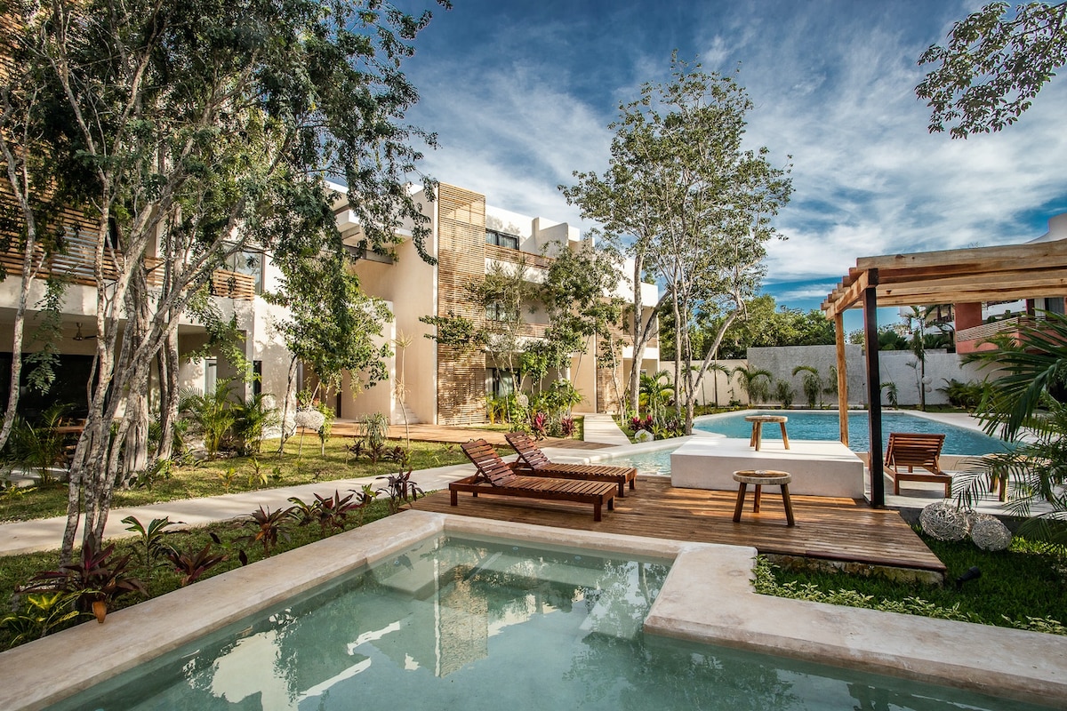 A serene common area is depicted, showcasing two large swimming pools surrounded by lush greenery. The shallow pool is positioned in the foreground with wooden decking, while sunchairs provide a relaxing space for guests. The vibrant landscape complements the tranquil atmosphere.