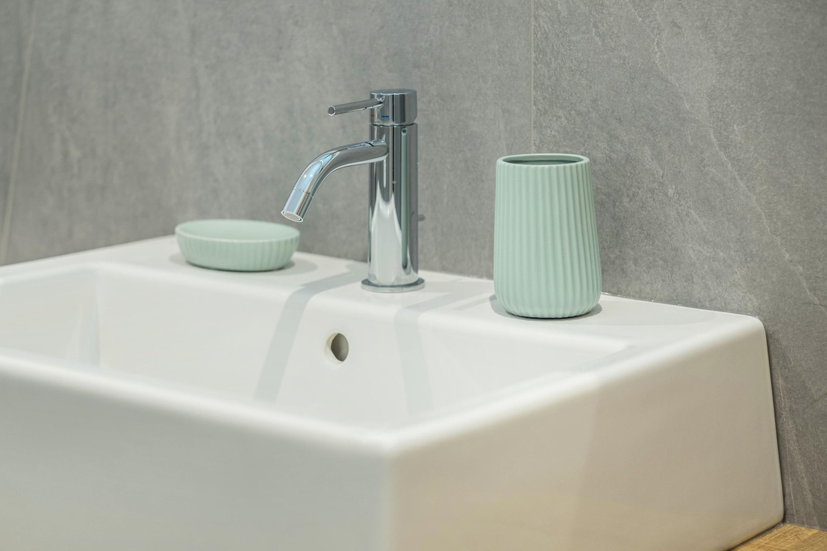 A modern bathroom sink is presented in the image, featuring a sleek, chrome faucet. Stylish mint-colored accessories, including a ribbed cup and a small dish, are positioned on either side of the sink, complemented by smooth, light grey tile walls.