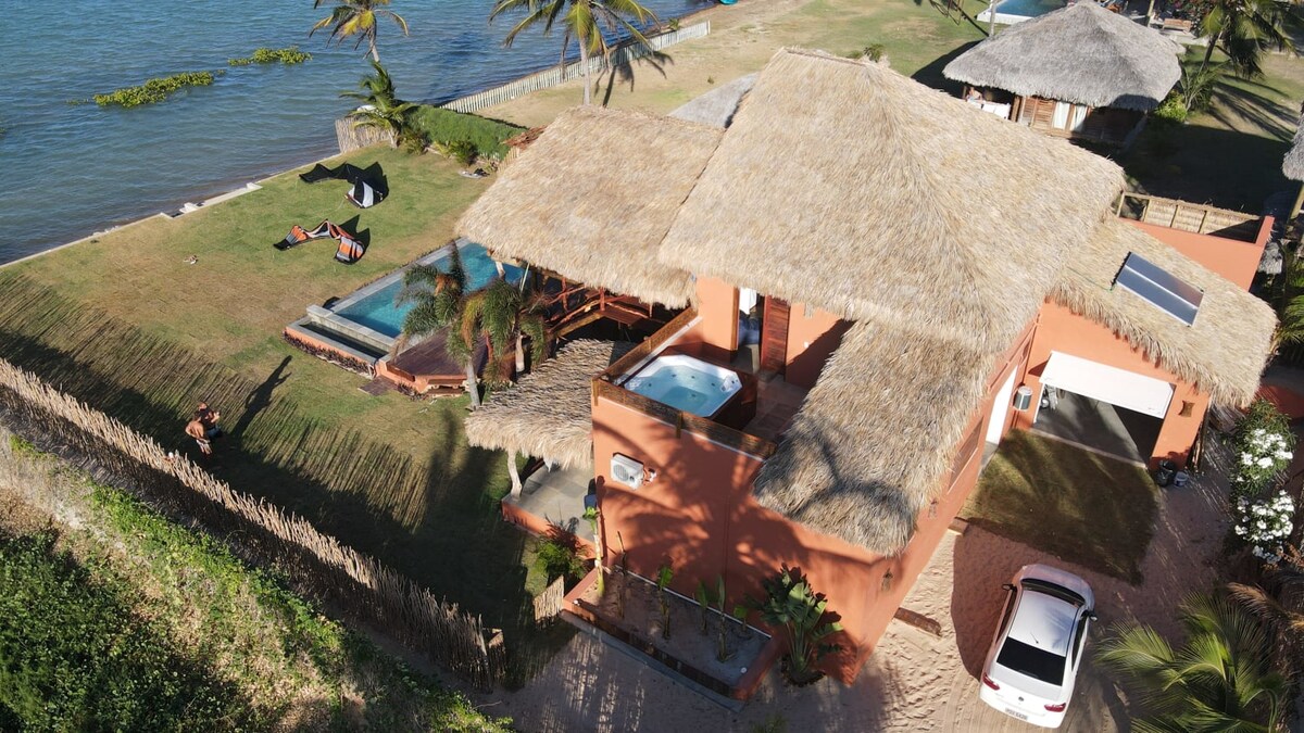The villa is situated by the water, surrounded by greenery and palm trees. A thatched roof provides shade over the main living area. An inviting pool is visible, along with lounge chairs positioned on the lawn, all enhancing the outdoor leisure space.