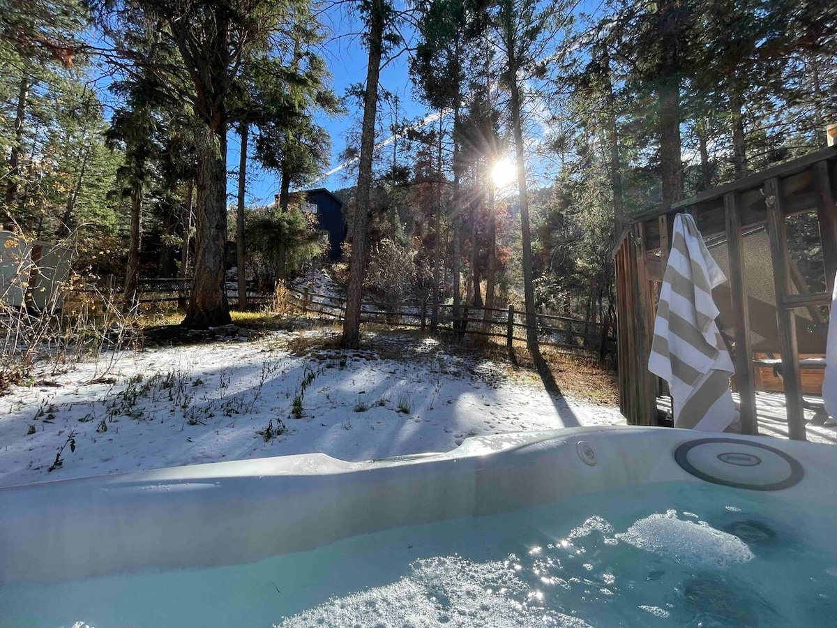 A hot tub is situated outdoors amidst a serene forest setting, surrounded by tall trees. Sunlight filters through the branches, illuminating sections of snow-covered ground nearby, while a striped towel hangs on the deck railing.