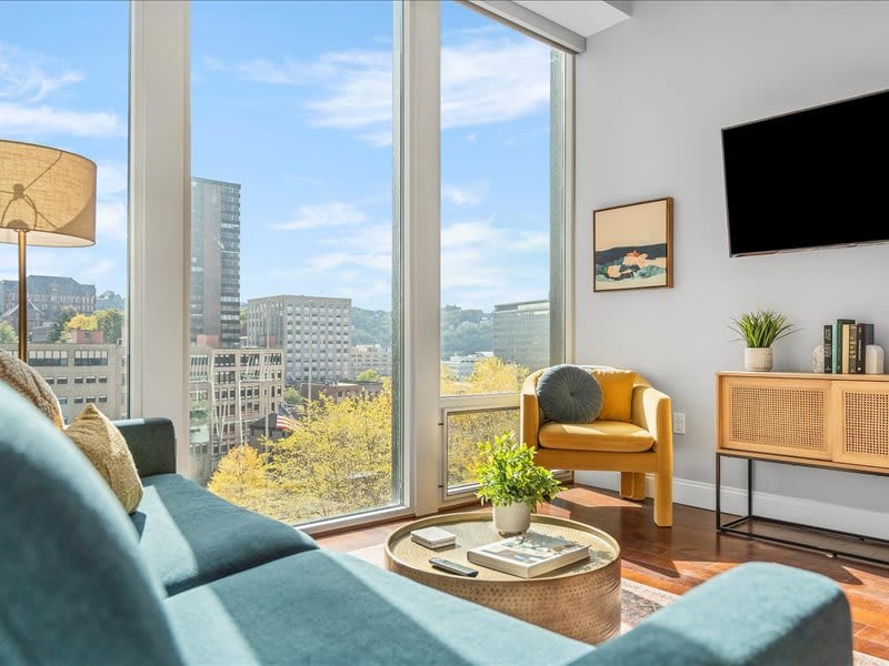 A bright living area features large windows showcasing city views. A cozy teal sofa contrasts with a mustard-yellow chair, while a circular coffee table holds decorative items. Natural light enhances the space, creating a welcoming environment for relaxation.