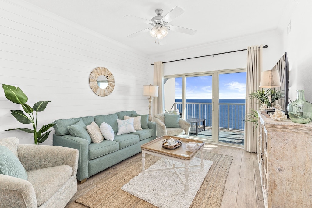 A bright living area features a light blue sofa and an armchair, complemented by a round mirror above a decorative console. Large sliding doors open to a balcony with views of the Gulf, while soft natural light enhances the warm wooden flooring.