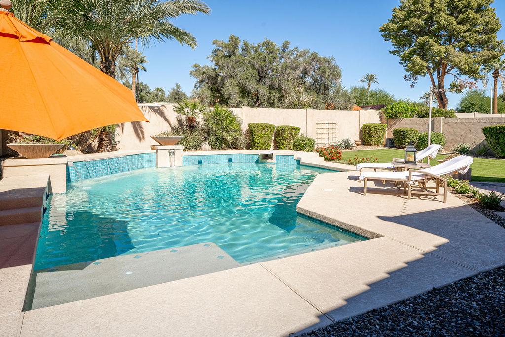 The outdoor area features a sparkling pool surrounded by well-manicured gardens, providing a tranquil oasis. An orange umbrella casts shade over comfortable lounge chairs, inviting relaxation in the Arizona sun. Lush greenery and vibrant landscaping enhance the serene atmosphere.