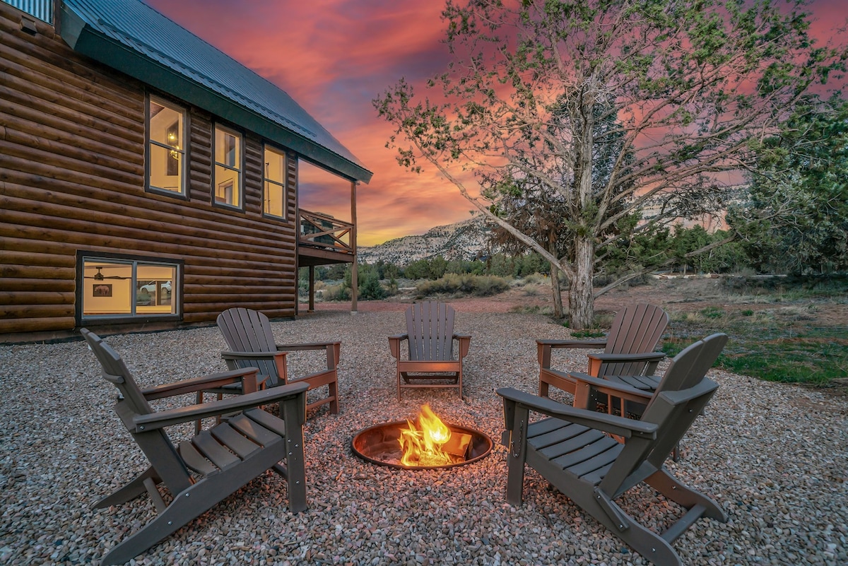 An outdoor fire pit is surrounded by four wooden Adirondack chairs, set on a pebbled area. In the background, the log cabin structure features large windows that reflect the vibrant hues of a sunset sky, enhancing the tranquil setting.