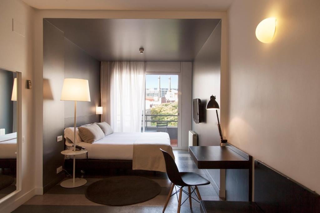 A spacious room is presented with a large window draped in sheer curtains, allowing natural light to illuminate the interior. A comfortable bed is positioned adjacent to a desk and chair. A floor lamp and wall-mounted light provide additional illumination.