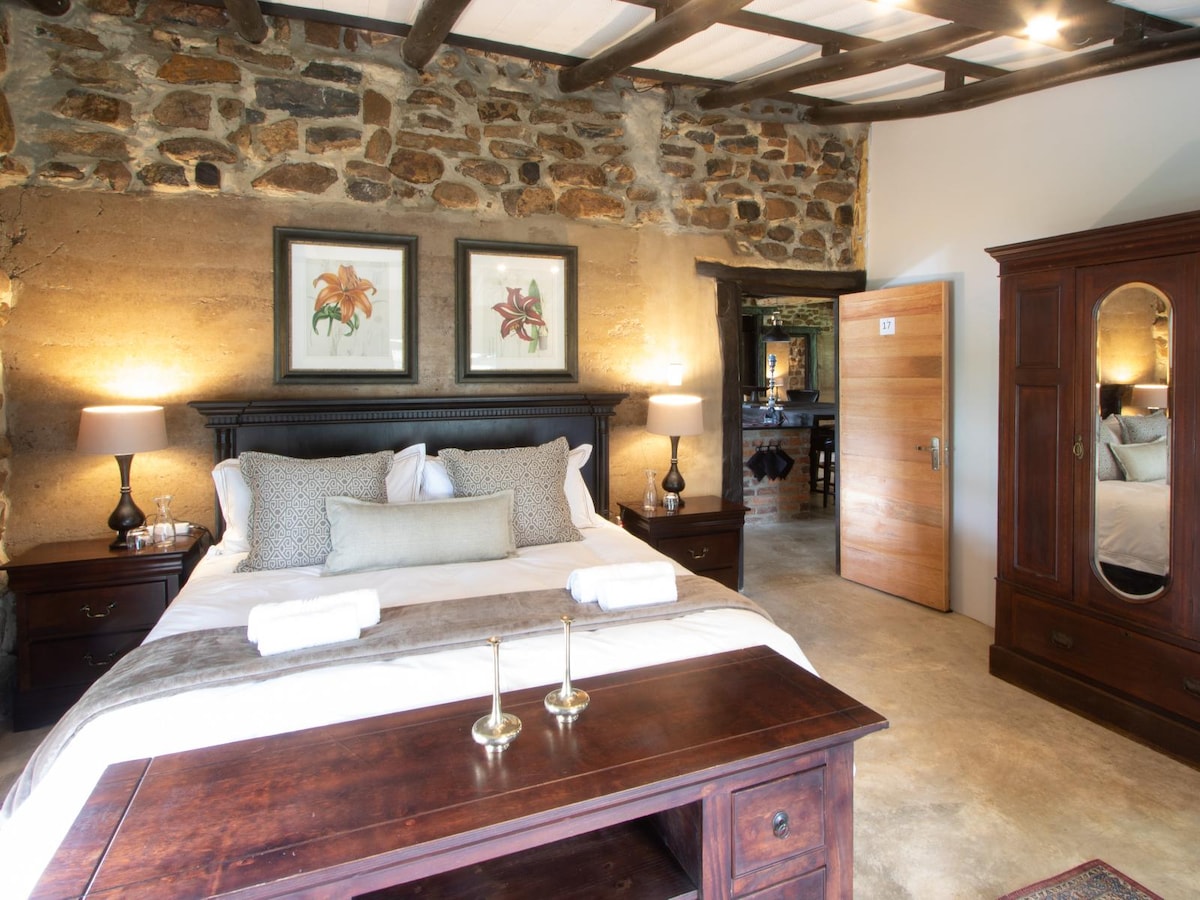 A spacious bedroom features a wooden bed framed by stone walls. Two framed floral artworks hang above, complementing bedside tables with lamps. A dresser with a mirror adds depth, while light illuminates the room’s natural textures and warm tones.