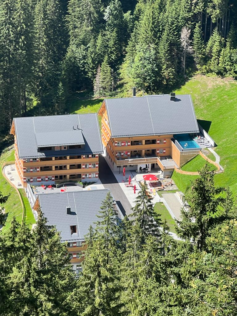 An aerial view captures a cluster of modern buildings nestled among greenery, featuring a swimming pool and landscaped grounds. The structures showcase a blend of wooden exteriors and balconies, while the surrounding trees create a natural backdrop.