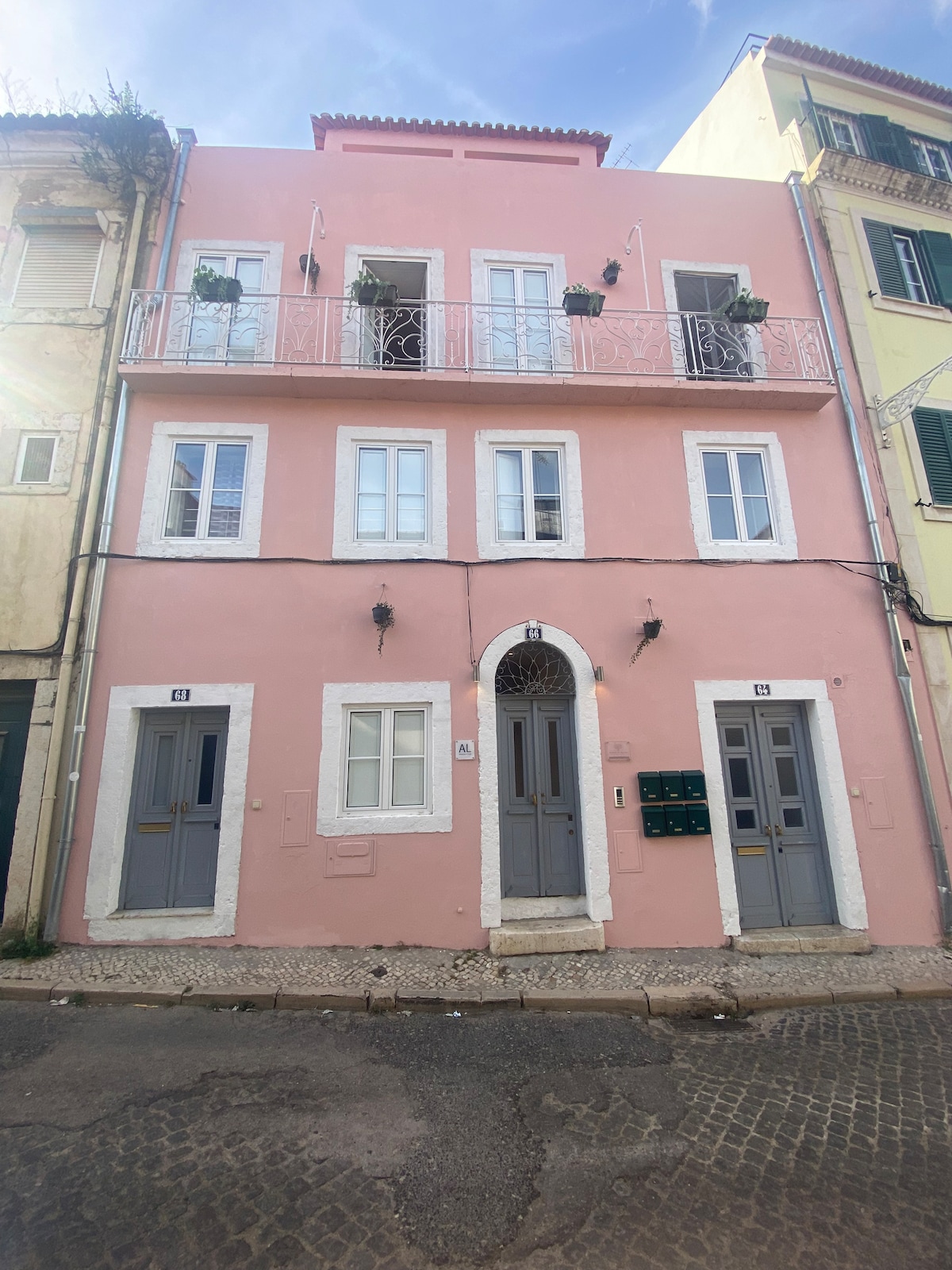 The building features a charming pink facade with decorative white trim and is accented by a small balcony adorned with plants. Dual gray doors serve as the main entrance, framed by elegant architectural details. Mailboxes are located beside the entrance.