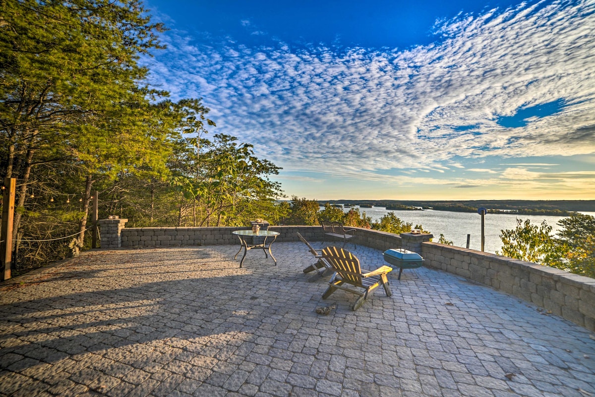 A spacious patio is illuminated by soft sunlight, featuring a set of wooden lounge chairs and a small dining table. The natural stone flooring complements the serene landscape, which overlooks Kentucky Lake and is framed by trees set against a backdrop of expansive, cloudy skies.