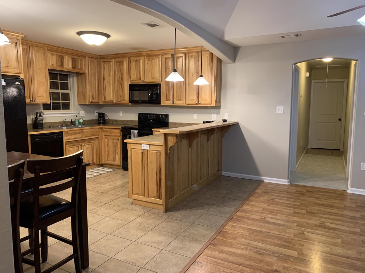 The kitchen showcases wooden cabinetry and modern appliances, including a black refrigerator and microwave. A breakfast bar is present, accompanied by four wooden stools. The tiled floor connects to a spacious living area, highlighted by natural lighting from overhead fixtures.
