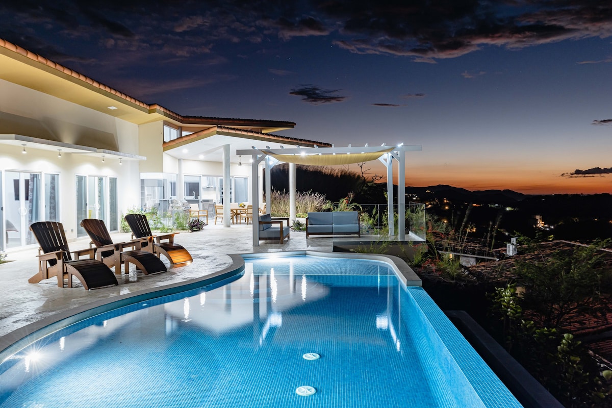 A custom-designed outdoor space features a modern pool illuminated by soft lighting, surrounded by comfortable seating and loungers. Large glass doors provide access to the interior, where the serene landscape views are complemented by the evening sky's hues.