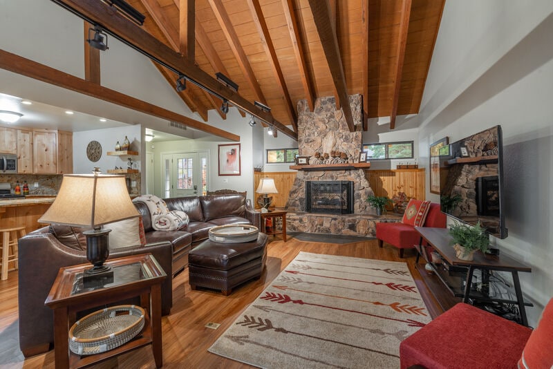 The living area features a leather sofa and two accent chairs, complemented by a coffee table and lamps. A stone fireplace is the focal point, set against a backdrop of exposed wooden beams. Natural light enters through windows, highlighting the rich wood flooring.