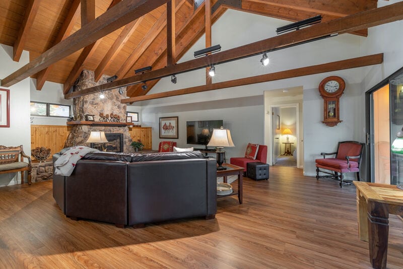 An inviting living room features a dark leather sectional sofa arranged around a coffee table. Wooden beams highlight the high ceiling, while a large stone fireplace serves as a focal point. A TV is mounted on the opposite wall, with soft lighting provided by table lamps.