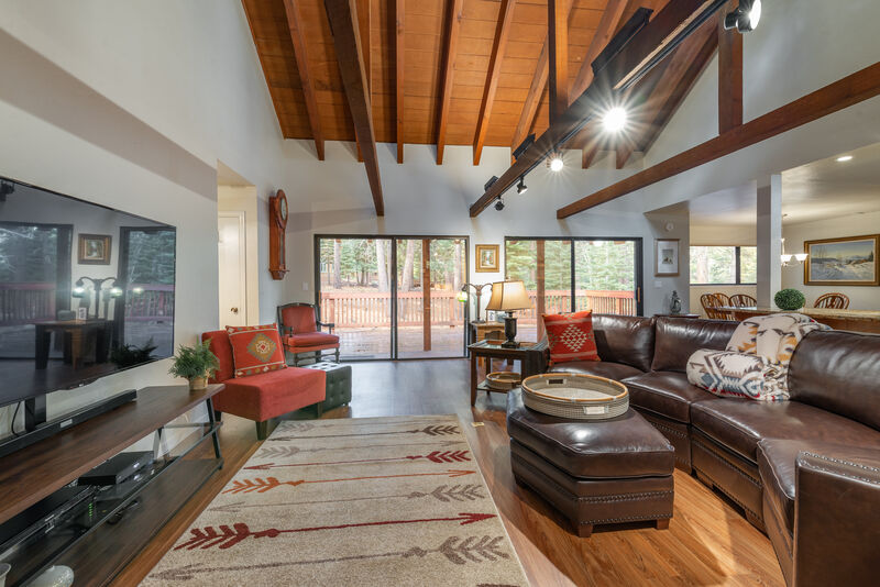 The spacious living room is highlighted by a vaulted ceiling with exposed wooden beams. A large leather sectional sofa is paired with a cozy armchair, while natural light floods in through sliding glass doors, leading to the outdoor deck. A patterned rug adds warmth to the wood floor.