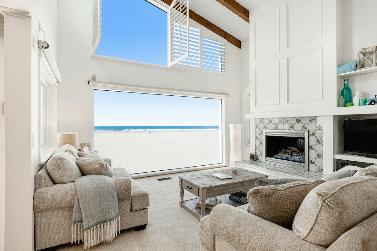 A spacious living area features large windows that frame expansive ocean views. A cozy seating arrangement includes light-colored sofas and a coffee table. A gas fireplace is positioned beneath the wall-mounted television, creating a focal point while allowing natural light to fill the room.