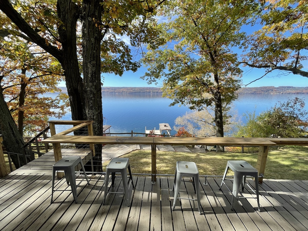 The expansive deck features bar height seating for four, overlooking a serene lakeside view. Surrounding trees display autumn colors, and the calm water reflects the blue sky. A grassy area leads down to the water, enhancing the tranquil atmosphere.