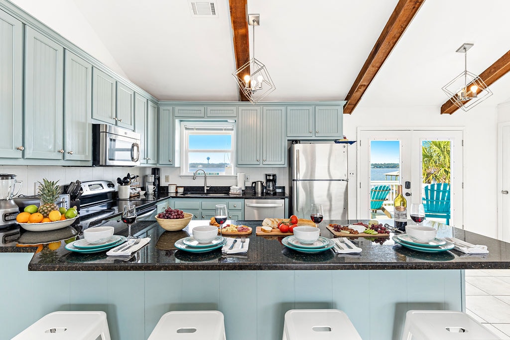 The kitchen features light blue cabinetry and granite countertops, complemented by wooden beams overhead. A large island is set with plates, glasses, and an assortment of fruits and snacks. Stainless steel appliances are visible, with double doors leading to an outdoor view.
