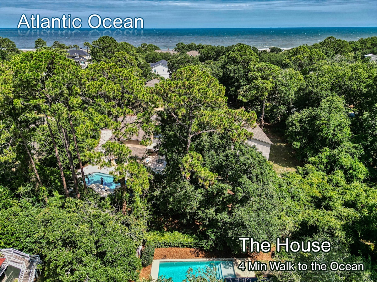 Aerial view of a beach house surrounded by trees, showcasing a large private pool in the backyard. The Atlantic Ocean is visible in the distance, with a path indicating a short walk to the beach. The lush greenery creates a serene atmosphere.