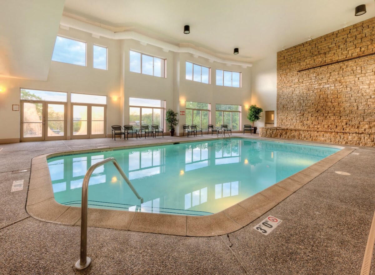 An indoor pool area is showcased, featuring large windows that allow natural light to fill the space. The pool is surrounded by a smooth deck, with seating available nearby. A stone wall adds texture to one side of the area, enhancing the inviting atmosphere.