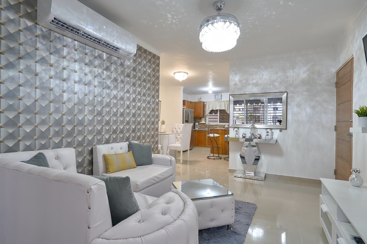 A modern living room is highlighted by elegant white furniture, including a pair of armchairs and a coffee table. A textured accent wall adds depth, while a sleek chandelier provides overhead lighting. The space flows into the dining area, visible in the background.
