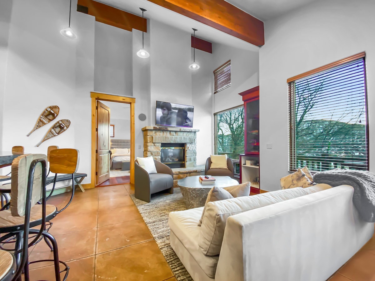 A living area is depicted with high ceilings and natural light streaming through windows. A stone fireplace is centered between two seating arrangements, featuring a cozy couch and armchairs. Rustic decor accents include snowshoes mounted on the wall, enhancing the mountain retreat atmosphere.