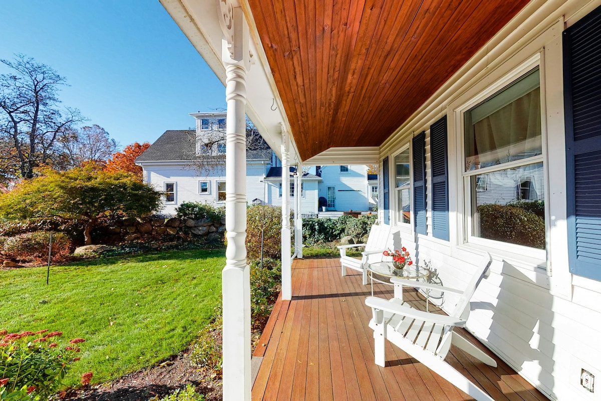 A covered porch features white rocking chairs set against a wooden floor. Green grass and landscaped plants surround the area, while a blue sky and sunlight enhance the inviting atmosphere. The home showcases a classic design with wood detailing above the entrance.