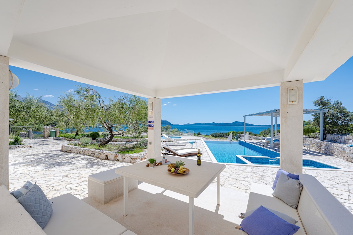An outdoor seating area is featured, with a shaded table surrounded by plush cushions. A clear view of the heated pool and the sea is visible in the background, complemented by landscaped greenery and olive trees, creating a serene and relaxing environment.
