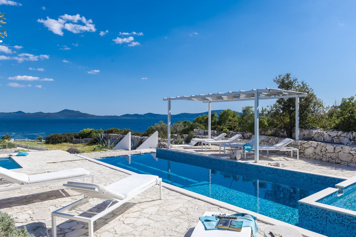 A sunlit terrace features a heated swimming pool surrounded by lounge chairs. A shaded pergola provides a relaxing space, with views of the sea and distant islands. The stone pathway leads to a landscaped garden filled with greenery, enhancing the outdoor setting.