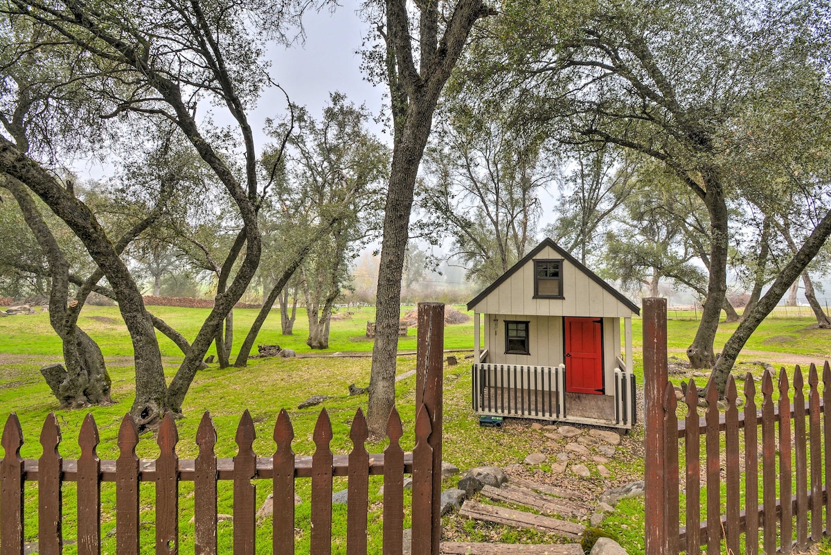 A charming playhouse is nestled among mature trees, featuring a red door and a welcoming front porch. The fenced yard is bordered by a wooden picket fence, enhancing the inviting outdoor space surrounded by lush greenery.