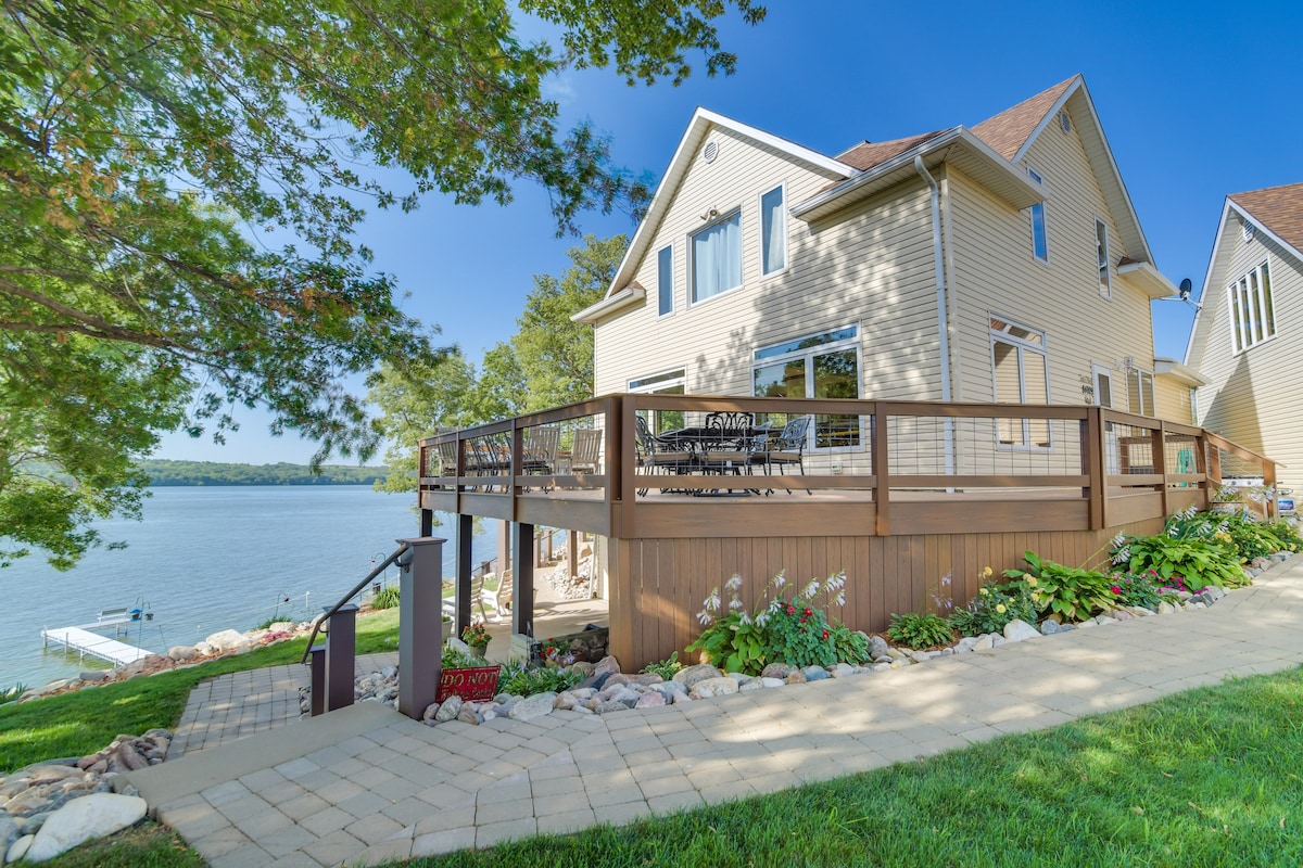 A spacious deck extends from the house, featuring multiple outdoor dining chairs surrounded by lush greenery and colorful flowers. Lake views are visible in the background, providing a serene waterside setting. The path leading down to the lake is bordered by landscaped stones.