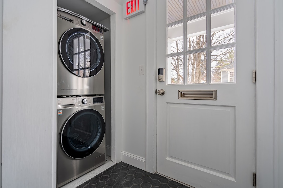 A compact laundry area features a stacked washer and dryer in sleek stainless steel, positioned near a door that leads outside. Natural light filters through the glass panes of the door, creating a bright and functional space.