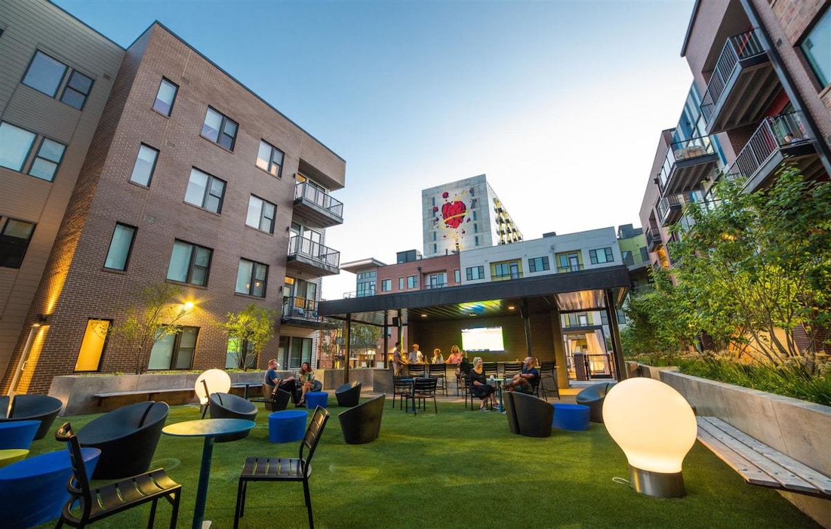 An outdoor lounge area features several round tables surrounded by colorful chairs on lush green grass. Soft lighting accentuates the space, with a large glowing orb sculpture and a pavilion. Residents are seen enjoying the area against a backdrop of modern apartment buildings.