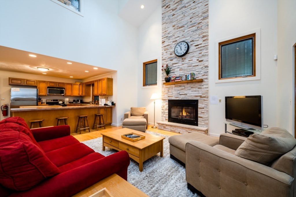 A spacious living area features a warm color palette with a large red sofa and a beige couch. A wooden coffee table sits in the center, and a stone fireplace is positioned against the wall, complemented by a wall clock and windows that allow natural light.