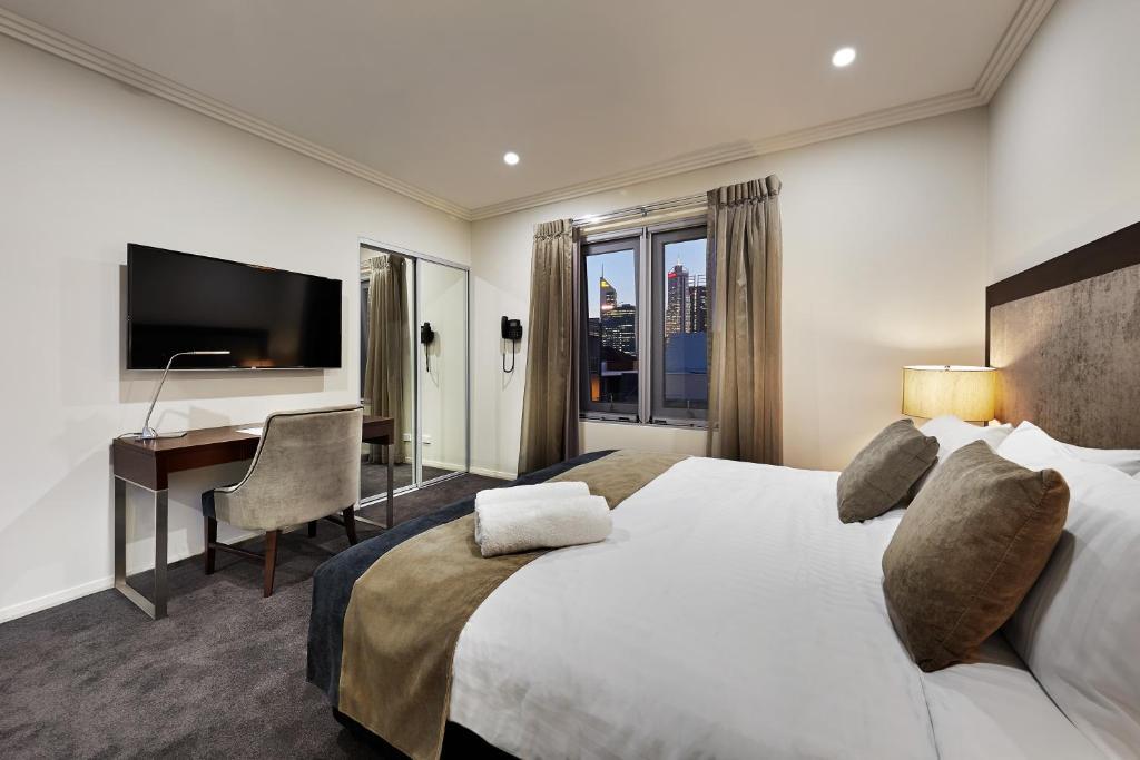 A bright and inviting bedroom features a spacious bed with neutral linens and decorative pillows. A sleek desk and chair are positioned near a large window dressed with soft curtains. A modern television is mounted on the wall, reflecting city views in the background.