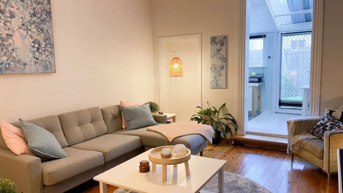 A cozy living area features a light gray sectional sofa, accented with soft pink pillows. A small white coffee table holds decorative items. Natural light flows in through a glass door leading to a private courtyard, creating a bright and inviting atmosphere.