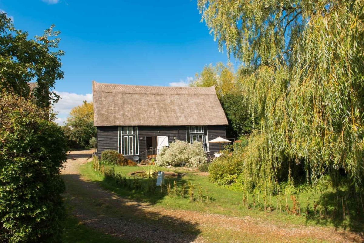 The thatched barn conversion is nestled in a green, rural setting, surrounded by well-maintained gardens and trees. A light blue sky enhances the peaceful atmosphere, with casual outdoor seating visible on the patio. The property is accessed via a gravel drive, leading to a welcoming entrance.