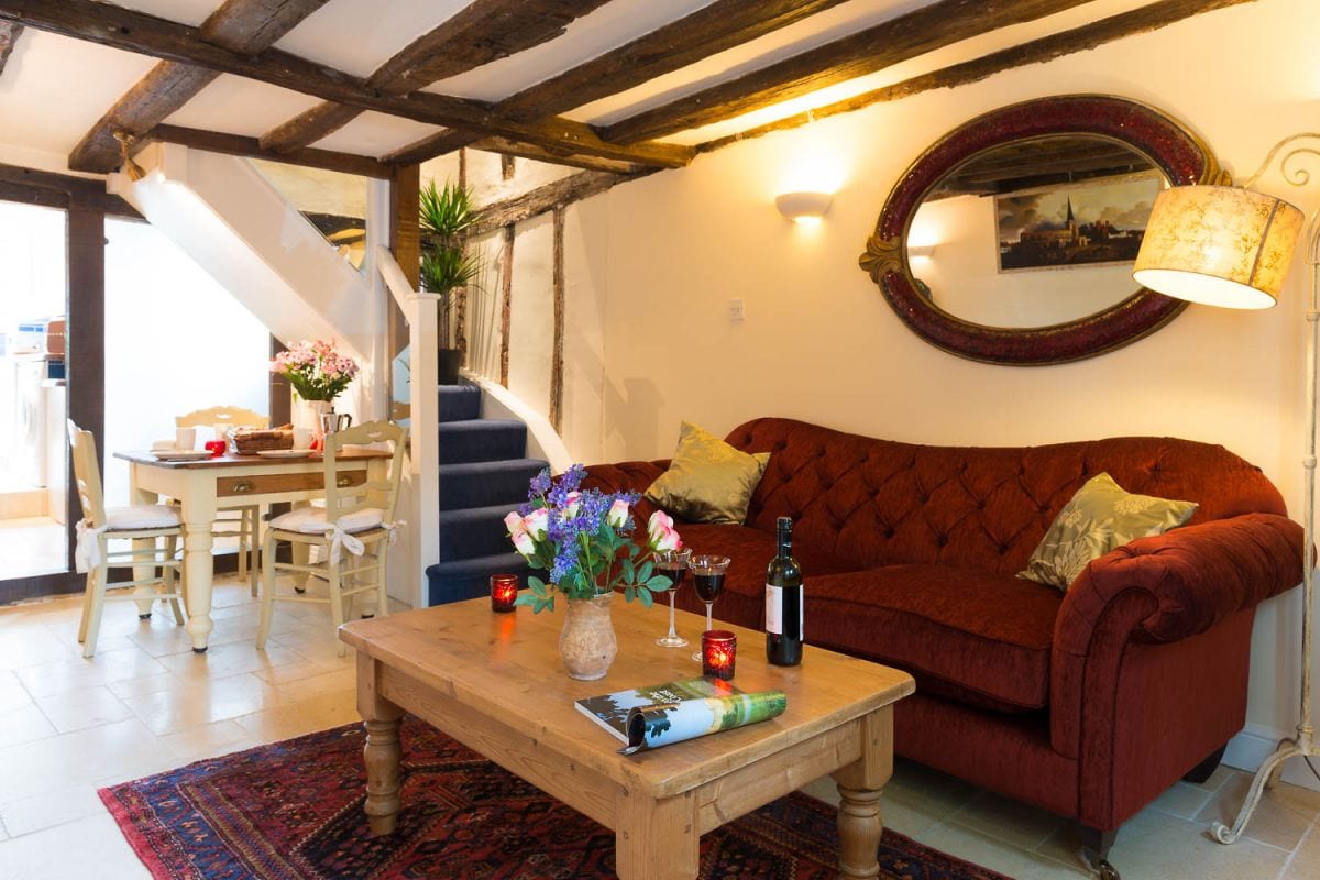 A cozy sitting room is highlighted by a rich red sofa and a wooden coffee table. An elegant mirror adorns the wall, reflecting a warm ambiance. Dining chairs are visible in the background, complementing the staircase with wooden beams that accentuate the historic charm of the cottage.