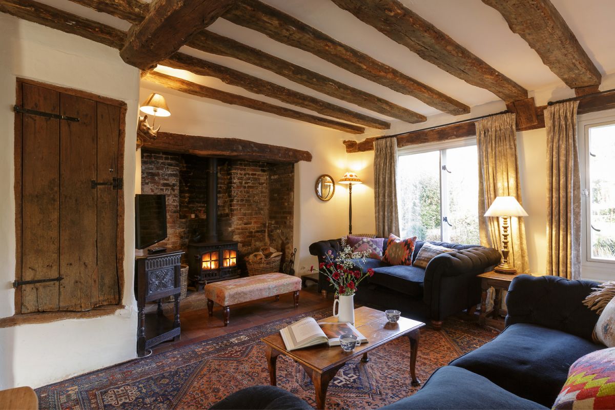 The inviting sitting room features exposed beam ceilings and a large inglenook fireplace with a cozy log burner. Two comfortable sofas are arranged around a wooden coffee table adorned with a book and a vase of flowers. Large windows offer views of the garden and allow natural light to fill the room.