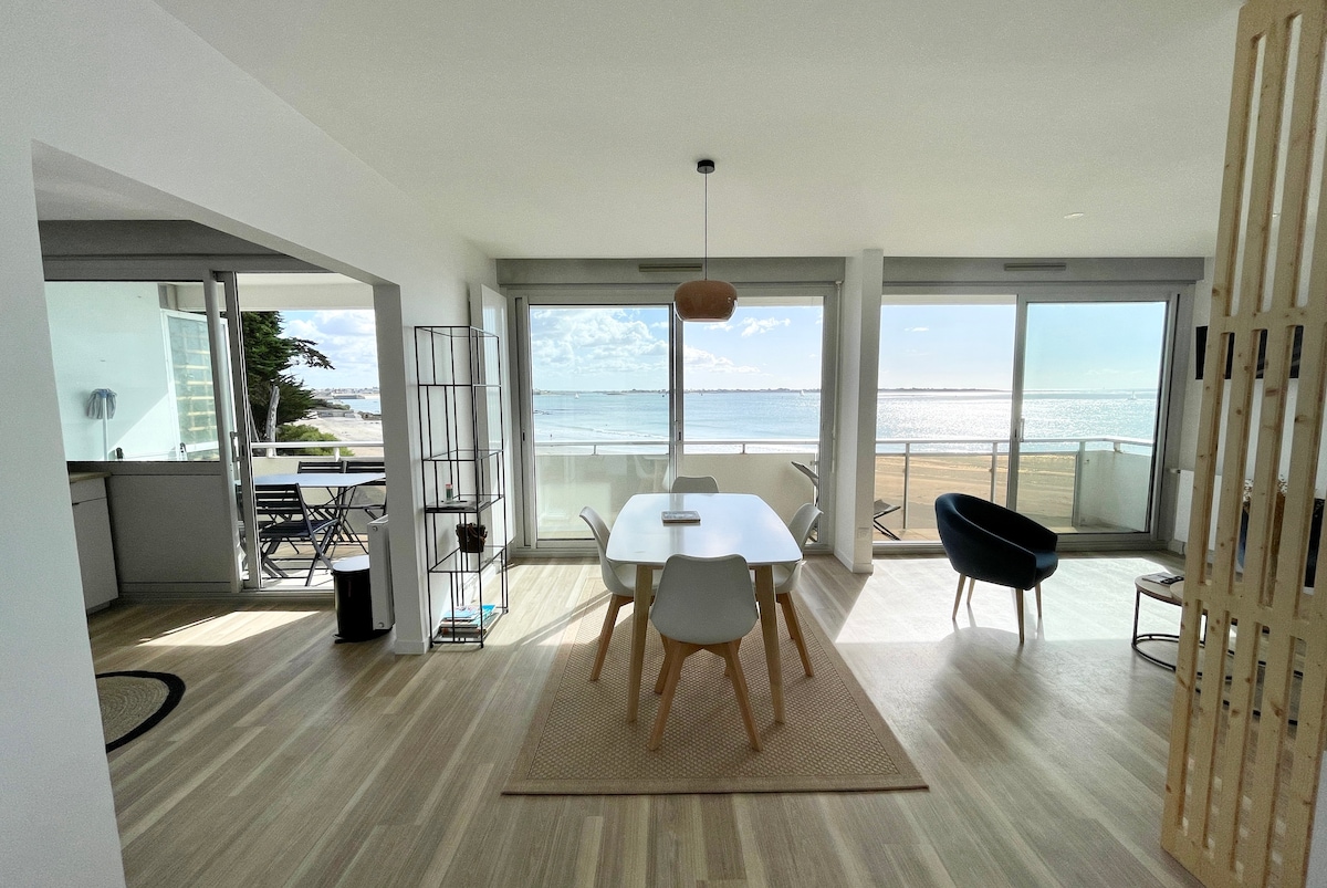 A luminous living area features a dining table with modern chairs, positioned beside expansive glass doors that reveal an ocean view. A cozy lounge chair and a textured area rug enhance the space, while the light wood flooring complements the natural elements.