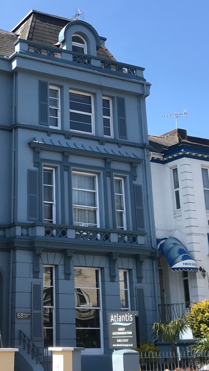 The exterior of the Atlantis guest house is presented, featuring a blue façade with detailed architectural accents. The building houses multiple stories. A sign indicating 'Atlantis' and 'Guest Accommodation' is visible at the entrance, complemented by a neighboring structure showcasing a traditional facade.