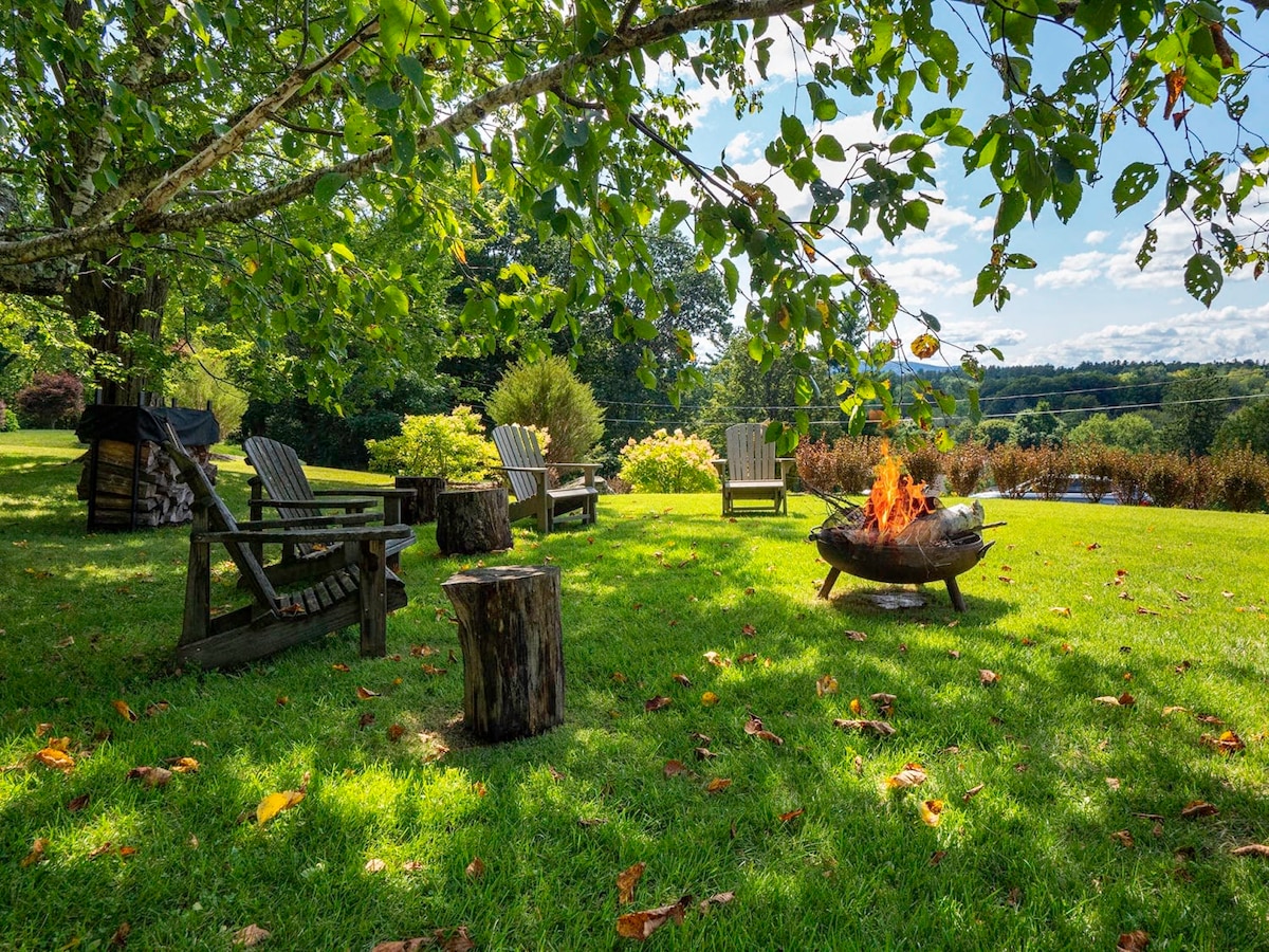 A spacious outdoor area is featured, surrounded by lush greenery and colorful landscaping. Multiple wooden chairs are arranged around a fire pit, with logs and nature providing a tranquil setting for relaxation. A picturesque view of rolling hills can be seen in the background.