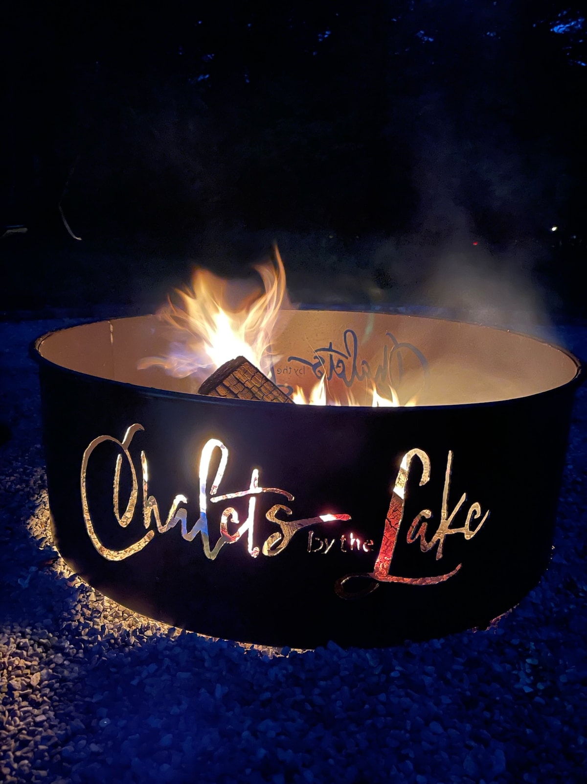 A fire pit is situated in a gravel area, featuring a decorative metal rim engraved with the words 'Chalets by the Lake.' Flames dance above the logs, creating a warm glow against the dark surroundings.