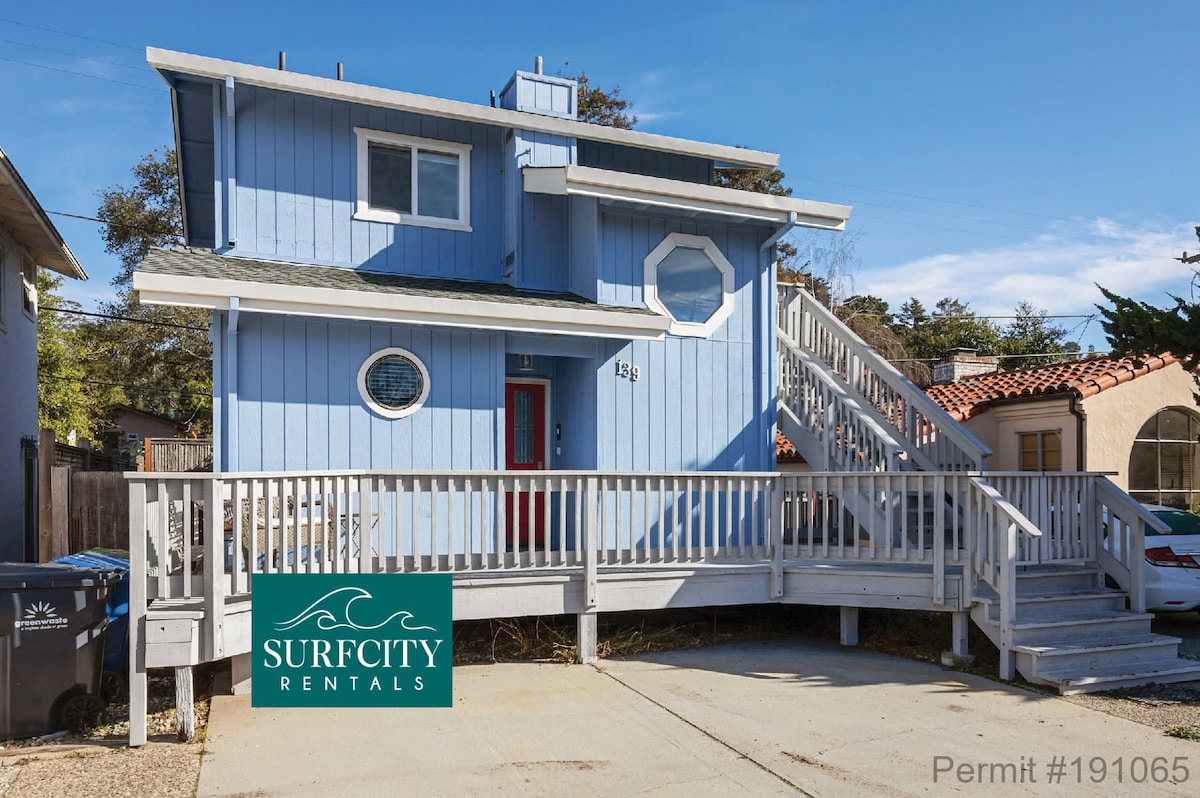 A charming beach house is shown from the exterior, featuring blue siding with white trim. A broad front deck with a ramp provides easy access, and distinctive circular windows enhance the facade. Surrounding areas include previous parking spots and a glimpse of nearby trees.